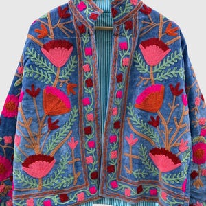 May include: A blue velvet jacket featuring vibrant floral embroidery in shades of pink, orange, and red. The garment has a high collar, long sleeves, and an open front design. The interior lining is visible, showcasing a light blue and white striped pattern.