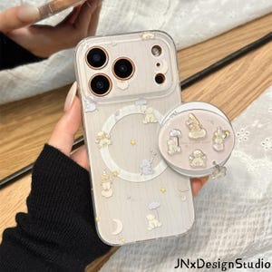 May include: A clear smartphone case featuring a pattern of small bears, clouds, stars, and moons in soft pastel tones. The case includes a circular magnetic ring and a matching circular phone grip decorated with several small bear illustrations.