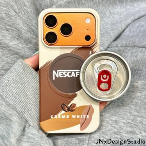 May include: A cream-colored smartphone case featuring a brown circular Nescafé logo, coffee bean graphics, and the text CRÈME WHITE. The case has an orange camera module and is paired with a circular silver phone grip with a red pull tab.
