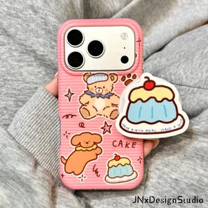 May include: A pink striped phone case featuring a cartoon bear, a dog, and two cake illustrations. A circular pop-out grip attached to the case displays a blue and yellow cake with the text HAVE A NICE MEAL HAVE A NICE CAKE printed on it.