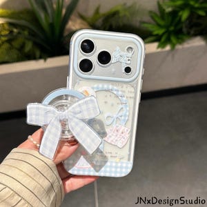 May include: A clear smartphone case with a light blue and white gingham pattern, featuring a matching gingham bow phone grip. The case includes a camera lens protector frame in light blue and various small decorative stickers with text.