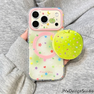 May include: A clear smartphone case with a pink camera frame and a pink circular magnetic ring. The case features a pattern of small colorful stars, hearts, and moons. A round, bright green apple-shaped phone grip with glittery star accents.