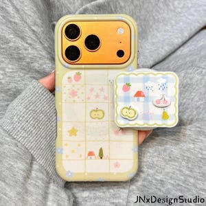 Può includere: Una custodia per smartphone giallo pallido con un motivo a griglia che presenta piccole icone di fragole, stelle, fiori, case e alberi. È fissato un supporto per telefono quadrato con bordi smerlati e stampa a quadretti blu e bianchi.