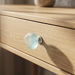 May include: A light blue, translucent, pebble-shaped glass knob attached to a light oak wood drawer. The knob features a smooth, rounded surface with a metallic silver-toned base that secures it to the front of the wooden furniture piece.