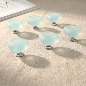 May include: A set of six cabinet knobs featuring smooth, frosted light blue glass tops with irregular, rounded shapes. Each knob is attached to a polished silver-toned metal base, creating a uniform look for furniture hardware and drawer pulls.