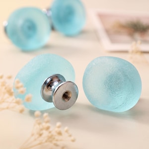 Puede incluir: Un juego de pomos para armarios de cristal esmerilado en color azul claro con forma redondeada similar a un guijarro. Cada pomo cuenta con una base de metal plateado pulido con un orificio central roscado para su fijación.