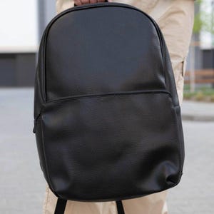 May include: A black backpack with a textured, pebbled surface. The bag features a large main compartment with a rounded top and a smaller, horizontal zippered front pocket. It has black shoulder straps and a top handle for carrying.