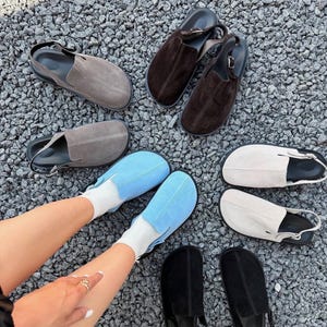 May include: A collection of suede slingback clogs in various colors including dark brown, light gray, beige, black, and bright blue. Each shoe features a rounded toe, a central seam, and an adjustable heel strap with a metal buckle closure.