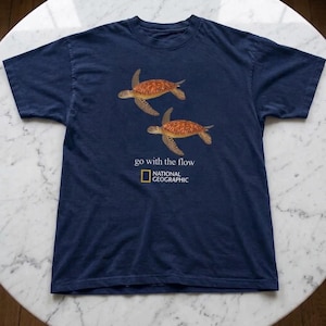 Puede incluir: Una camiseta azul marino de manga corta con dos tortugas marinas naranjas nadando sobre el texto "go with the flow" y el logotipo de National Geographic en blanco y amarillo. La prenda tiene cuello redondo y un corte estándar.
