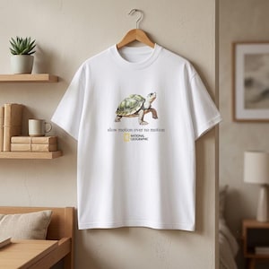 Puede incluir: Una camiseta blanca de manga corta con un gráfico de una tortuga en el centro. Debajo de la tortuga, el texto dice "slow motion over no motion" seguido del logotipo de National Geographic en negro y amarillo.