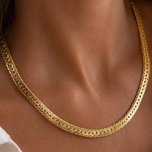 Peut inclure: Un collier chaîne doré au design à maillons chevrons plats et tressés. Le collier présente une surface polie et réfléchissante, reposant près du cou, mettant en valeur un motif continu et complexe de segments métalliques imbriqués.