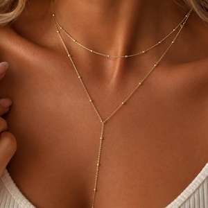 May include: A gold-toned layered necklace featuring two delicate chains adorned with small, evenly spaced spherical beads. The upper chain forms a simple choker style, while the lower chain extends into a Y-shaped drop design with a central vertical strand.