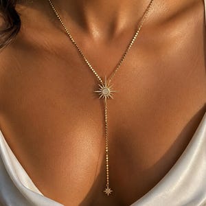 May include: A gold-toned lariat necklace featuring a central starburst pendant with a circular centre and radiating rays, accented with small clear stones. The chain consists of flat, reflective circular links, ending in a smaller starburst charm.