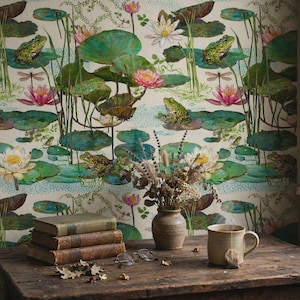 May include: Wallpaper featuring a repeating pattern of green frogs, pink and white water lilies, and lily pads on a cream background. A stack of three books, a ceramic vase with dried flowers, and a ceramic mug sit on a wooden table.