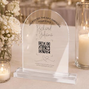 May include: An arched clear acrylic sign with a rectangular base featuring black printed text that reads HELP US SEE THE DAY THROUGH YOUR EYES, Richard + Juliana, and UPLOAD YOUR PICTURES TO OUR DIGITAL WEDDING GALLERY, with a QR code and heart.