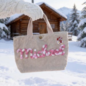 Può includere: Una borsa tote in velluto a coste beige con il nome Michael ricamato in lettere di filato a righe rosa e bianche. La borsa presenta una piccola toppa ricamata a forma di pupazzo di neve in alto a destra e una chiusura a bottone dorata.
