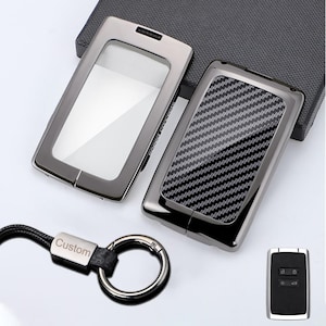 Può includere: Due cover rettangolari per chiavi auto con cornici grigio metallizzato. Una cover presenta una finestra trasparente, l'altra un motivo in fibra di carbonio nero e grigio. È incluso un portachiavi in pelle nera con anello in metallo argentato.