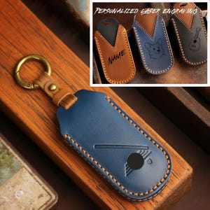 Può includere: Una custodia in pelle blu per chiavi con un anello in ottone e un cinturino in pelle color cuoio. La custodia presenta una grafica in rilievo e un ritaglio circolare. Un riquadro mostra tre custodie in pelle aggiuntive in cuoio, blu e nero con incisioni laser.