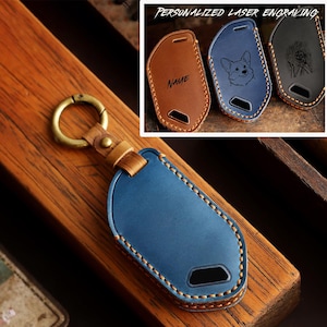 May include: A blue leather key fob cover with contrast stitching and a brass ring. The text PERSONALIZED LASER ENGRAVING appears above three additional covers in tan, blue, and black, featuring custom engravings of a name, a dog, and a ball.