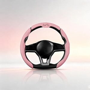 May include: A steering wheel cover featuring a textured light pink material with black accents. The top section of the pink cover displays the cursive text Elizabeth Taylor. The steering wheel has a black centre hub and a three-spoke design.