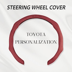 May include: A dark red steering wheel cover with a textured surface. The text STEERING WHEEL COVER is at the top, while TOYOTA PERSONALIZATION is in the center. The phrase LIVE DRIVE THRIVE is embossed on the upper inner rim of the cover.