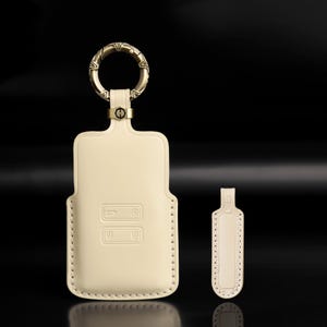 Può includere: Una custodia per chiave auto in pelle color crema dotata di un anello circolare in ottone anticato con incisioni geometriche e un piccolo passante in metallo con logo H. Un'etichetta rettangolare in pelle abbinata con bordi in rilievo è posta accanto.