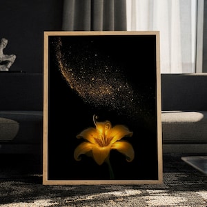 Könnte beinhalten: Ein rechteckiger Wandkunstdruck in einem hellen Holzrahmen, der eine einzelne leuchtende goldene Lilienblüte zeigt, die unten vor einem tiefschwarzen Hintergrund zentriert ist, mit einer Spur aus schimmernden Goldstaubpartikeln.