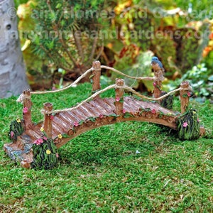 May include: A miniature wooden bridge with a curved design, featuring a textured surface and decorative floral accents. The bridge has rope railings and is adorned with a small blue bird figurine. The bridge is set on a bed of green moss.