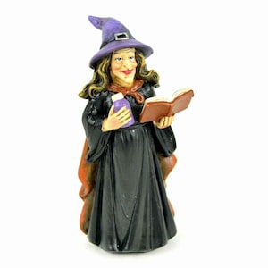 May include: A figurine of a witch wearing a purple hat and black dress, holding a purple bottle and a brown book. The witch has long, wavy hair and a red cape. A Halloween-themed decorative item.