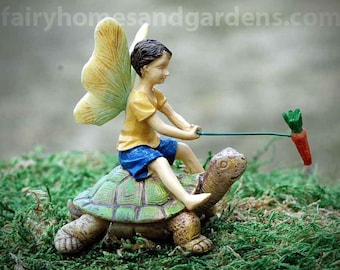 May include: A miniature fairy figurine with wings, wearing a yellow shirt and blue shorts, riding a green turtle with a carrot in its mouth. The fairy is holding a stick with a green leaf on the end.