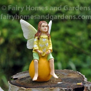 May include: A fairy figurine with wings, wearing a yellow and green dress, sits on a yellow bouncy ball. The fairy has brown hair and blue eyes. The figurine is on a tree stump.