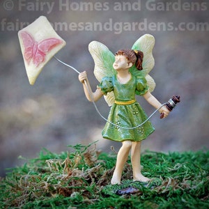 May include: A miniature fairy figurine with green wings and a green dress is holding a string attached to a pink and white kite. The fairy is standing on a bed of green moss.
