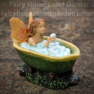 May include: A miniature fairy figurine is taking a bath in a green leaf-shaped bathtub. The fairy has orange wings and is holding a white pearl. The bathtub is filled with white bubbles and is sitting on a green base.