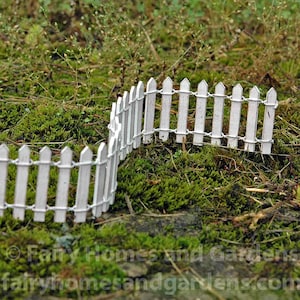 Könnte beinhalten: Zwei Abschnitte eines Miniatur-Weißzäuns, verbunden mit Draht, sind in einem Bett aus grünem Moos und Gras angeordnet. Der Zaun hat spitze Spitzen und ist für einen Feengarten oder eine Miniaturlandschaft konzipiert.