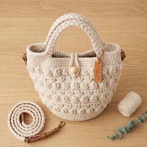 Puede incluir: Un bolso tipo cubo de ganchillo beige con un patrón texturizado de puntos de burbuja, un asa trenzada y un cierre central de cuenta de madera. Incluye una correa trenzada desmontable con detalles de cuero marrón y una etiqueta de cuero CROCHET & CO.