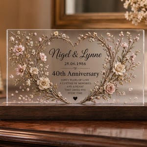 Può includere: Una targa rettangolare in acrilico trasparente su base in legno scuro, con una ghirlanda stampata a forma di cuore di fiori rosa e bianchi. Il testo recita: Nigel & Lynne, 28.06.1986, 40th Anniversary, Forty years of love, a lifetime of memories and a heart that chose you every time.