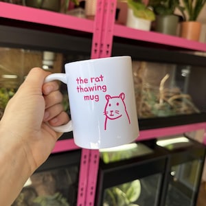 May include: A white ceramic mug featuring a pink graphic of a rat and the text "the rat thawing mug" printed in pink. The mug has a standard handle on the side and a smooth, cylindrical body. The design is centred on the front of the mug.