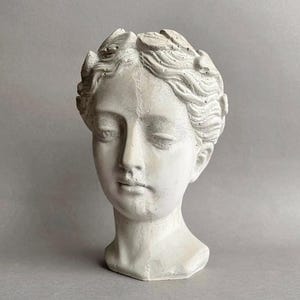 Puede incluir: Un busto de hormigón gris claro de una cabeza humana con una superficie texturizada y una corona de hojas que rodea el cabello. La escultura muestra una expresión facial serena con los ojos cerrados, una nariz recta y labios definidos.