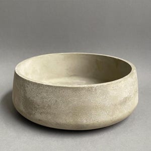 Könnte beinhalten: Eine runde, flache Schale aus hellgrauem Beton. Das Gefäß hat eine glatte, matte Oberfläche mit subtilen Textur- und Farbvariationen auf der Außenseite und zeichnet sich durch ein schlichtes, offenes Design mit einem dicken, festen Rand aus.