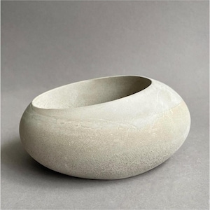May include: A light gray, rounded stone bowl with a smooth, matte texture and an asymmetrical, slanted opening. The vessel features a natural, porous surface appearance with subtle variations in tone, creating a simple, organic, and solid form.