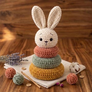 May include: A crochet stacking toy featuring a cream-colored rabbit head with long ears atop three plush rings in muted terracotta, sage green, and mustard yellow. The toy is accompanied by small yarn balls, a wooden crochet hook, and buttons.