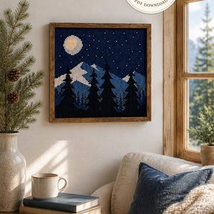 Puede incluir: Un bordado de punto de cruz cuadrado en un marco de madera que representa un paisaje nocturno con luna llena, cielo azul oscuro con estrellas, siluetas de pinos y montañas nevadas. La imagen incluye un logotipo circular con el texto DOMUS ARS FILUM.