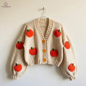 Könnte beinhalten: Eine beigefarbene Strickjacke mit V-Ausschnitt, langen Ärmeln mit gerippten Bündchen und einer Knopfleiste mit drei orangefarbenen Knöpfen. Das Kleidungsstück ist mit mehreren gehäkelten orangefarbenen Fruchtmotiven mit grünen Stielen verziert.