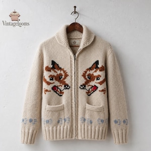 May include: A cream-colored knit zip-up cardigan featuring a shawl collar and two front pockets. The sweater displays two symmetrical fox head motifs in brown, black, and red, with light blue paw print patterns along the ribbed hem and cuffs.