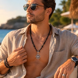 May include: A necklace featuring a round silver compass pendant suspended from a strand of small black beads. The pendant displays a detailed star design. The necklace is worn with a beige linen button-down shirt, a black beaded bracelet, and a wristwatch.