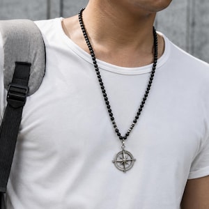 May include: A long necklace featuring small, smooth black spherical beads strung together, accented by a few metallic silver-toned beads near the center. A circular silver-toned pendant shaped like a compass with cardinal directions hangs below.