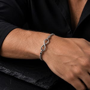 May include: A silver-toned metal bracelet featuring a central infinity symbol charm. The bracelet is constructed with a textured, flexible chain link design that connects to the infinity loop, creating a continuous and sleek appearance on the wrist.