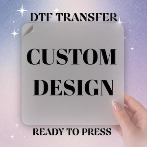 May include: A square DTF transfer sheet with rounded corners featuring the black text DTF TRANSFER, CUSTOM DESIGN, and READY TO PRESS. The sheet is a solid light gray color, presented as a flat, opaque material ready for heat application.