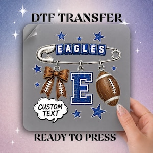 May include: A DTF transfer sheet featuring a silver safety pin with blue letter blocks spelling EAGLES, a brown bow, a large blue letter E, and a brown football. Includes a white speech bubble with the text CUSTOM TEXT and blue stars.