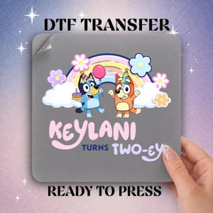 May include: A square DTF transfer sheet featuring a cartoon graphic of two blue and orange dogs wearing party hats, standing under a rainbow with clouds and flowers. The text reads KEYLANI TURNS TWO-EY. The design is on a gray background.